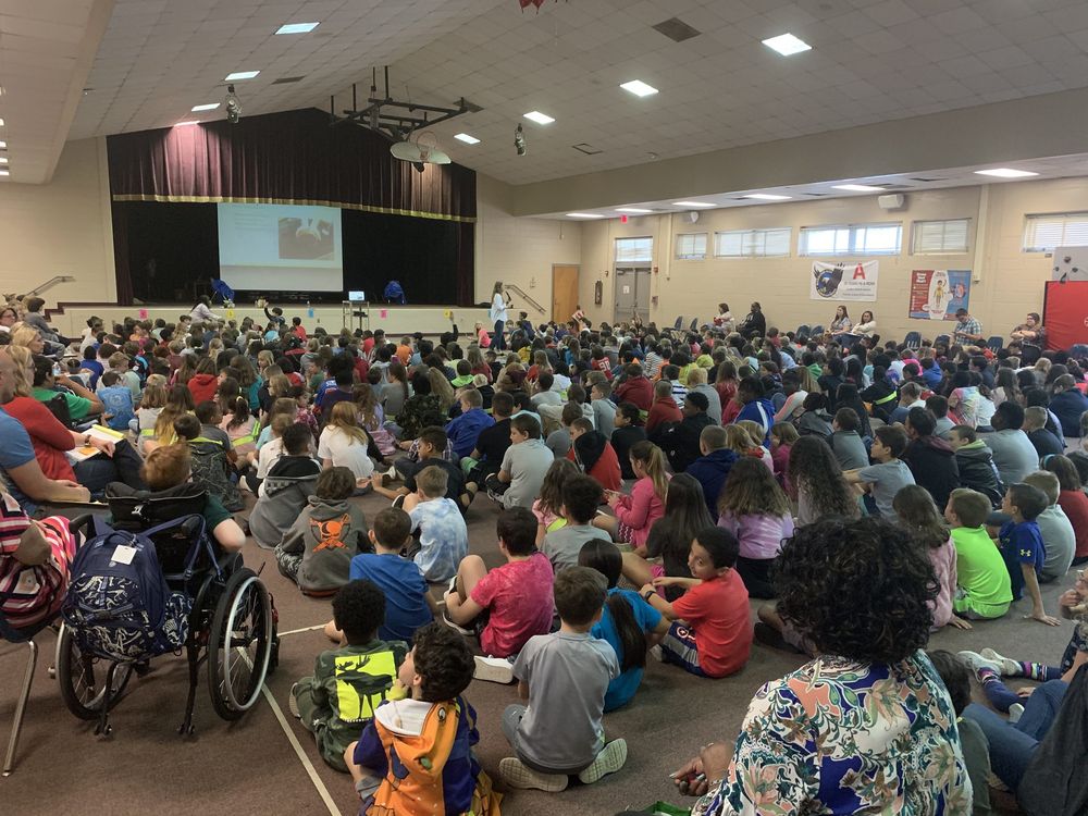 Students seated on a school gymnasium floor, facing a presentation on the stage.