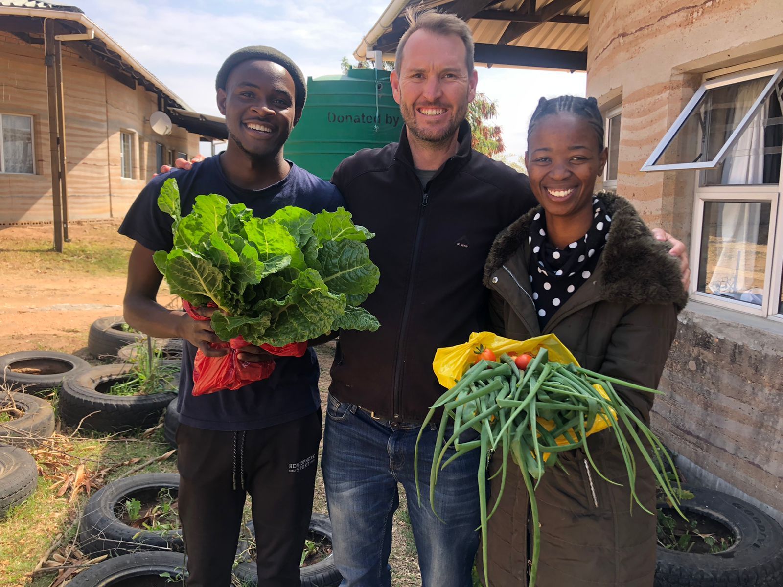 Three growers holding a fresh harvest of greens, spring onions and tomatoes outside a community greenhouse
