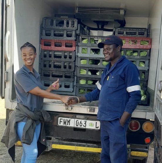 A grower and a driver shake hands at the back of a delivery truck stacked with crates of fresh produce.