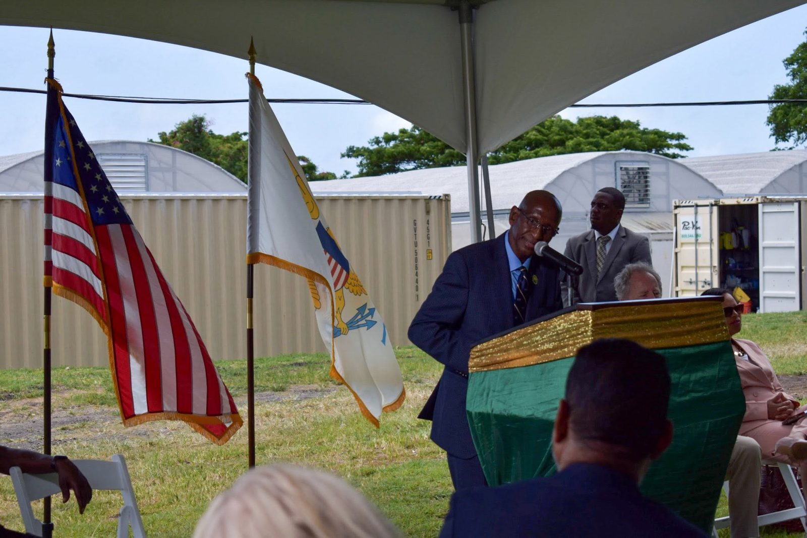 A speaker at a podium under a tent, the United States and Virgin Islands flags beside him and greenhouses behind.