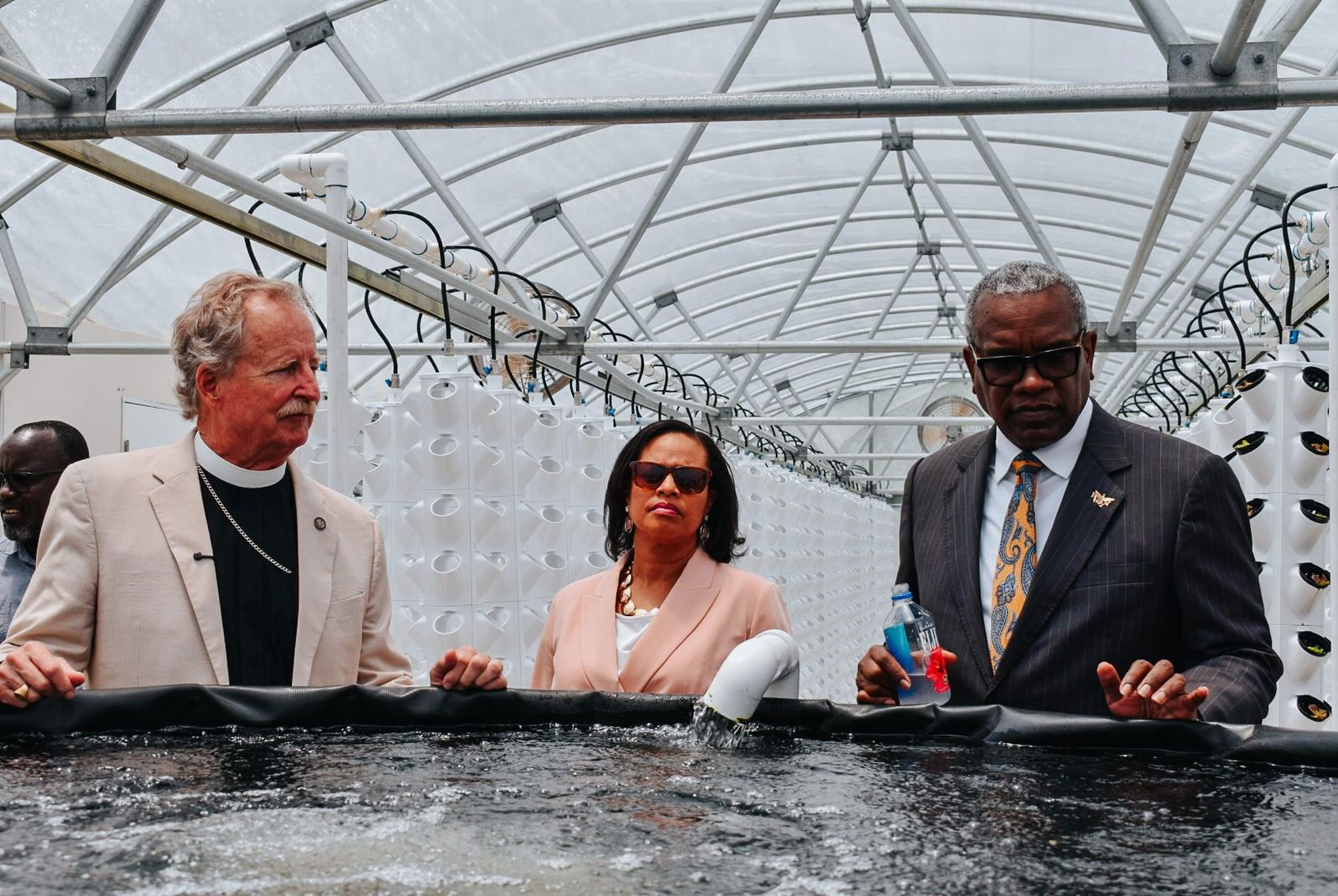 Founder Rev. Dr. Robert V. Lee III and two guests stand at an aerated fish tank inside an Island Fresh greenhouse, white tower-culture columns behind them.