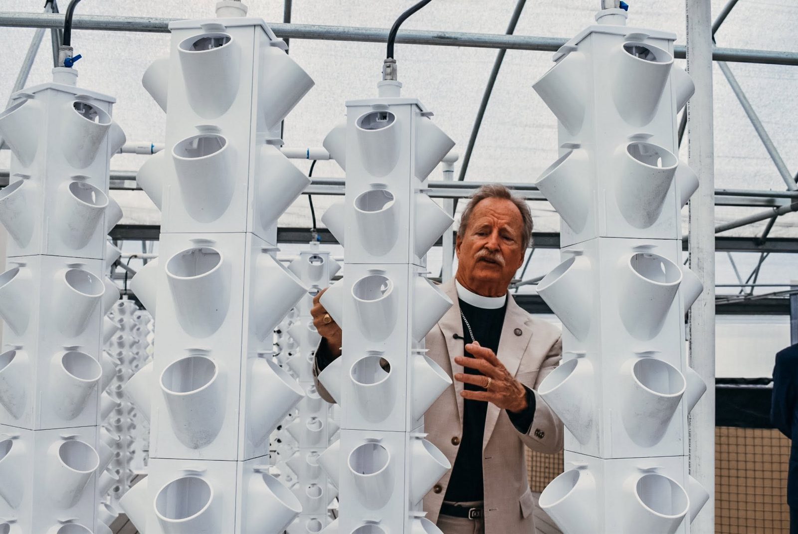 A man in a clergy collar speaks between tall white tower-culture growing columns inside a greenhouse.
