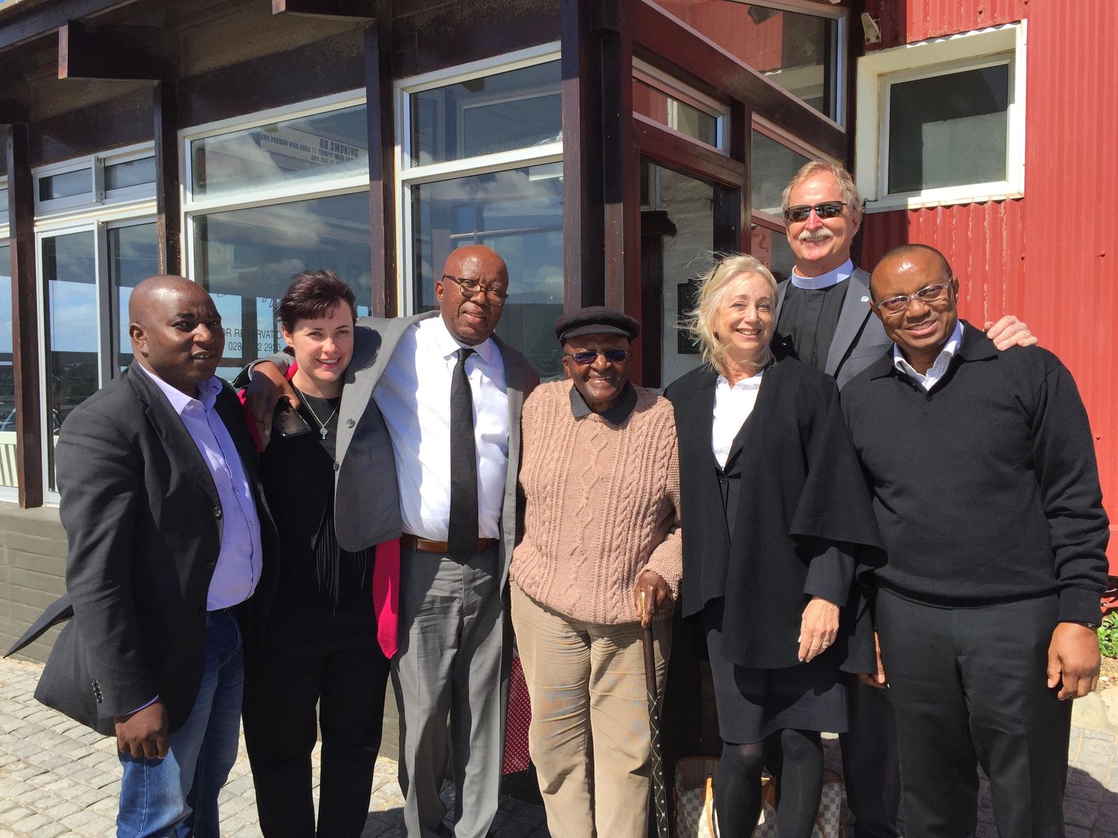 Archbishop Desmond Tutu stands arm in arm with FreshMinistries leadership and partners outside a red building in South Africa.