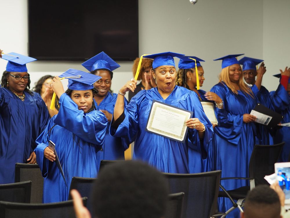 Graduates in blue caps and gowns celebrating at a FreshMinistries ceremony, one holding her certificate high.
