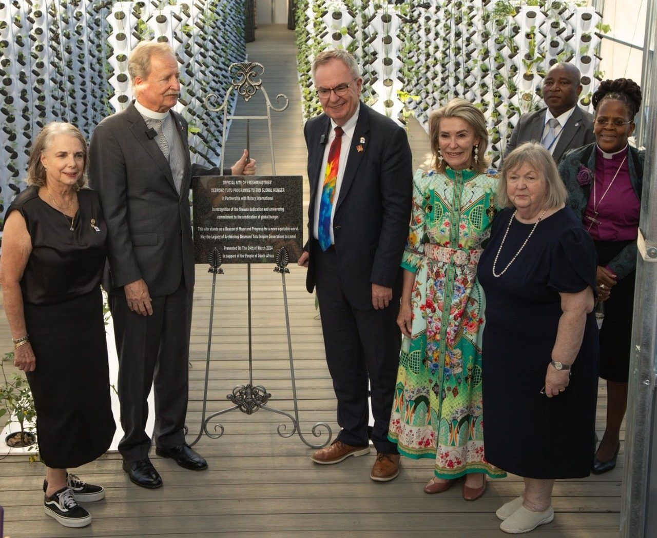 FreshMinistries leaders and partners stand beside a dedication plaque between rows of grow towers inside a tower greenhouse.