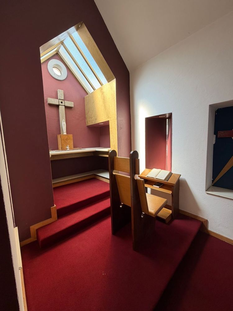 A small prayer room in the Tutu home: a stone cross on a deep red wall under a skylight, with a wooden prayer desk and an open book on a lectern.
