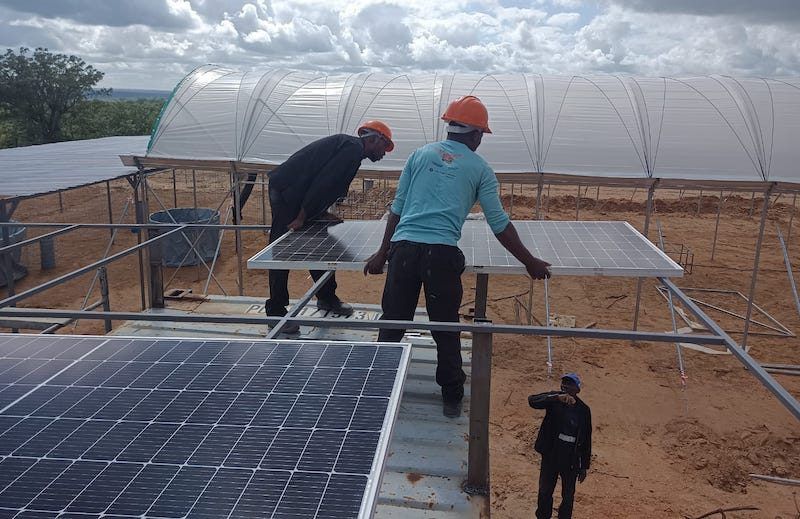 Workers in hard hats install solar panels on a frame beside a greenhouse under construction